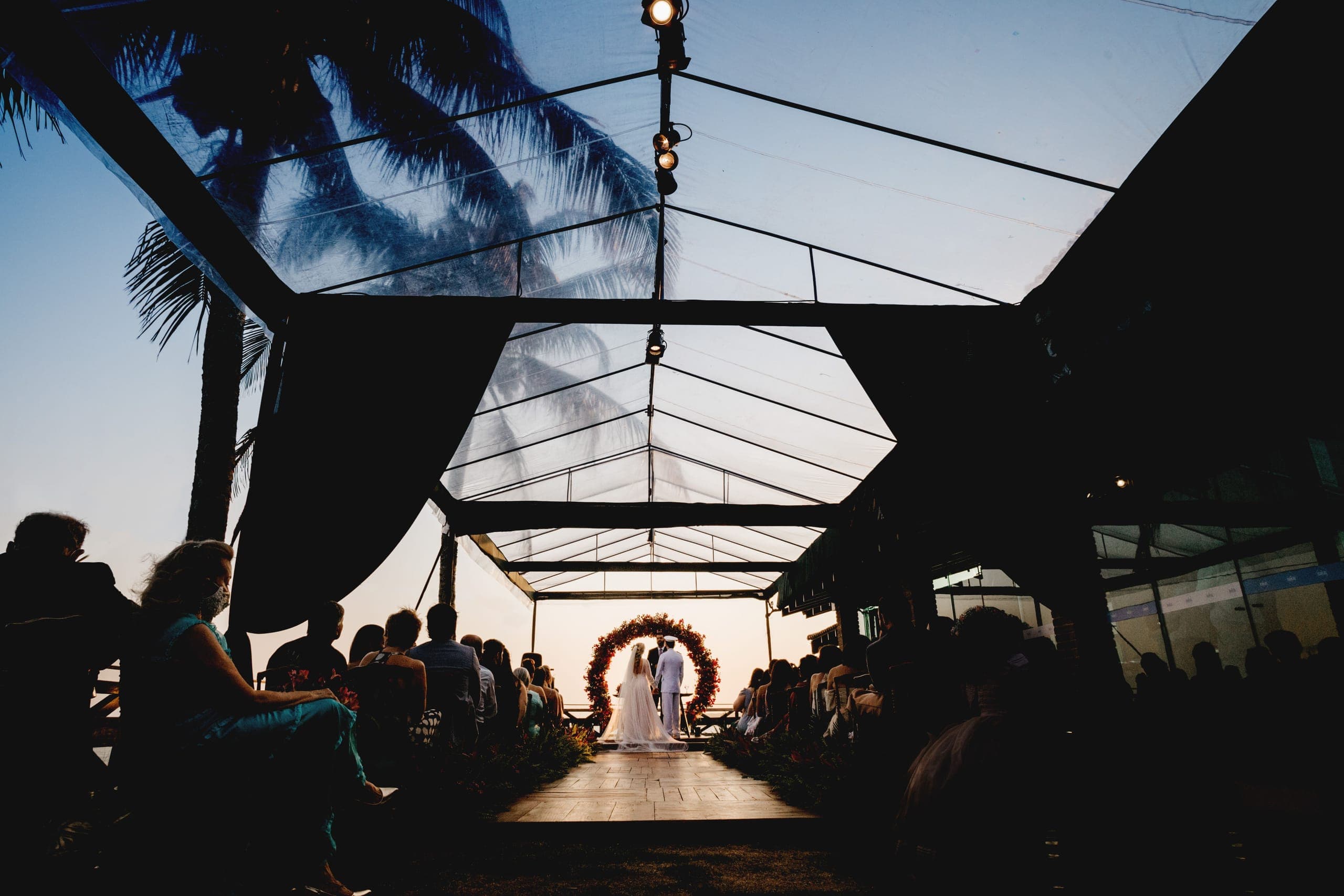 Cerimônia de casamento ao pôr do sol, corredor sob tenda com arco de flores